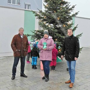 Zwei Männer und eine Frau mit einem Julius-Plüschtier stehen draußen vor einem großen Tannenbaum, um den sich Kinder vom Julius Kindergarten versammelt haben