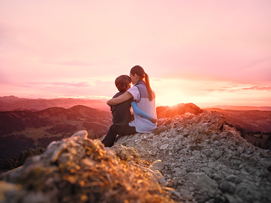 A mother sits on a mountain peak with her child in her arms and watches the sunset.