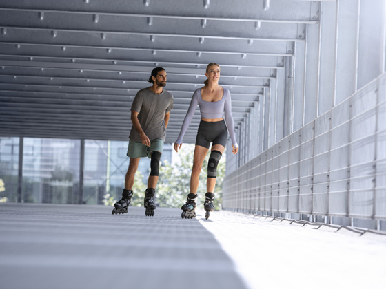 A man and a woman wearing knee braces are rollerblading in a garage.