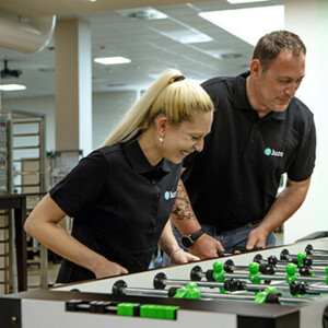 Eine Frau und ein Mann mit schwarzen Juzo T-Shirts spielen in ihrer Mittagspause mit Eifer Kicker