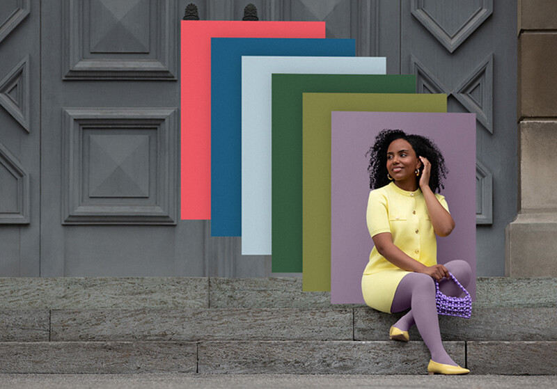 Juzo Trend Colours Batique Une femme est assise sur des marches en pierre devant un portail. Elle porte un ensemble deux pièces jaune clair et un bandage compressif violet. Derrière elle, on aperçoit des aplats de couleurs tendance.