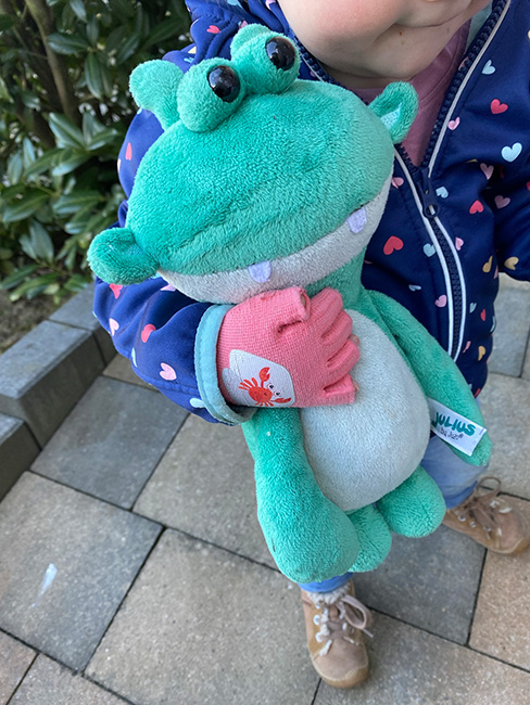 ein kleines Mädchen trägt an der Hand einen rosanen Kompressionshandschuh und hält ihr Julius Kuscheltier fest