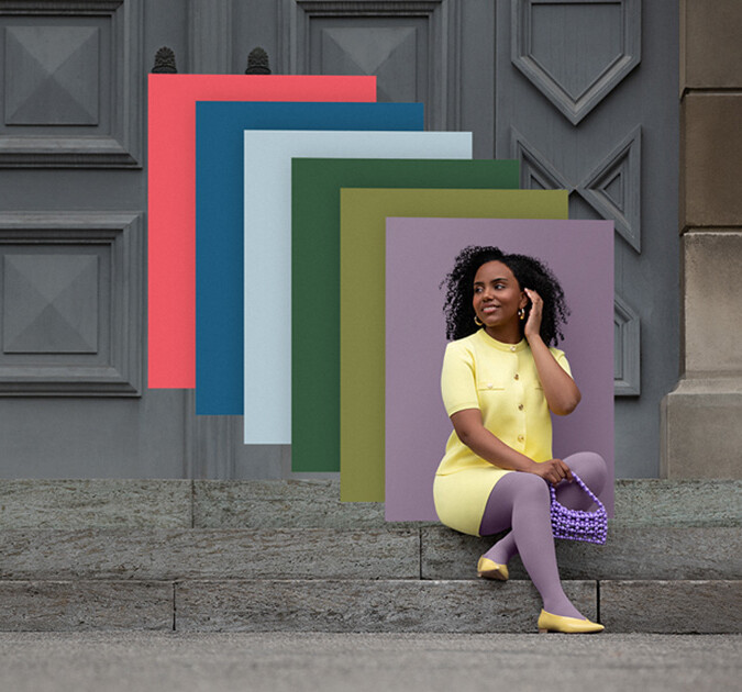 Juzo Trend Colours Batik Une femme est assise sur des marches en pierre devant un portail. Elle porte un ensemble deux pièces jaune clair et un bandage compressif violet. Derrière elle, on aperçoit des aplats de couleurs tendance.