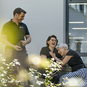 Zwei Frauen mit einem schwarzen Juzo T-Shirt sitzen auf einer Bank und lachen herzhaft, sie unterhalten sich mit einem Mann, der ebenfalls ein schwarzes Juzo T-Shirt trägt und eine schwarze Tasse in der Hand hält