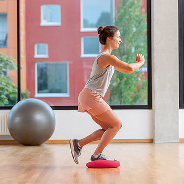 Uebung einbeinige Squats Schritt 2 Frau steht mit einem Bein auf einem weichen Kissten und geht in die Kniebeuge