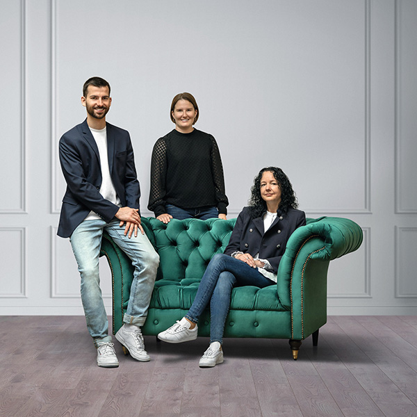 Auf dem Bild sieht man 2 Frauen und einen Mann, welche sich um ein grünes Sofa positioniert haben