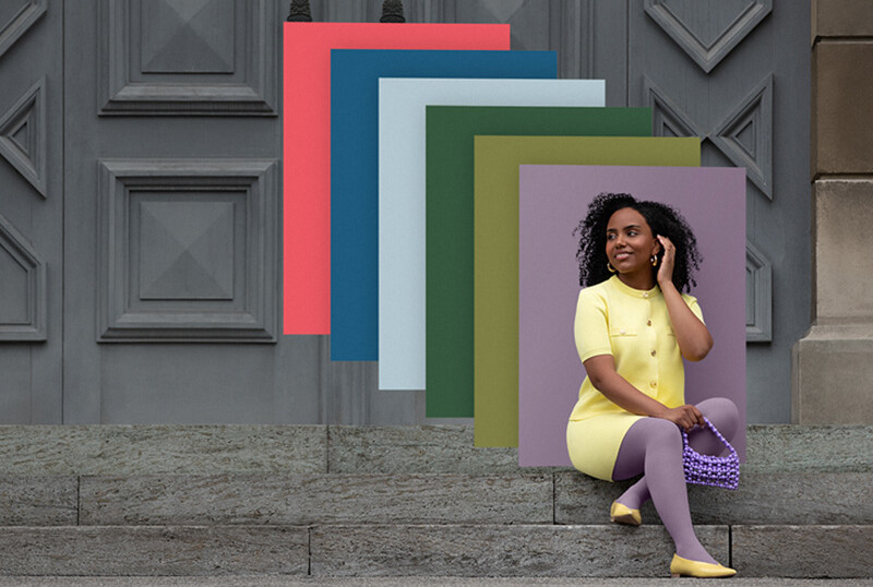 Juzo Trend Colours Batik Eine Frau sitzt auf Steinstufen vor einem Tor. Sie trägt einen hellgelben Zweiteiler zu einer lilanen Kompressionsversorgung. Hinter ihr sieht man Farbfelder in den Trend Colours