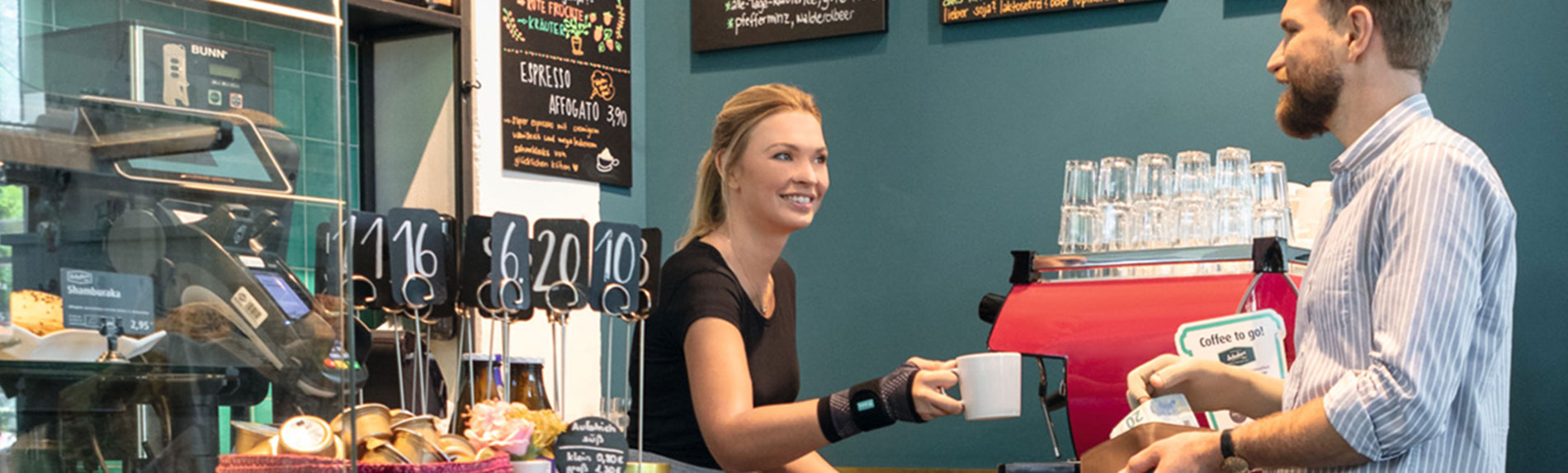 Lifestyle image - Cafe - JuzoFlex Manu Xtra Bakery employee wearing JuzoFlex Manu Xtra wrist support hands cup to customer over the counter