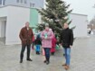 Zwei Männer und eine Frau mit einem Julius-Plüschtier stehen draußen vor einem großen Tannenbaum, um den sich Kinder vom Julius Kindergarten versammelt haben