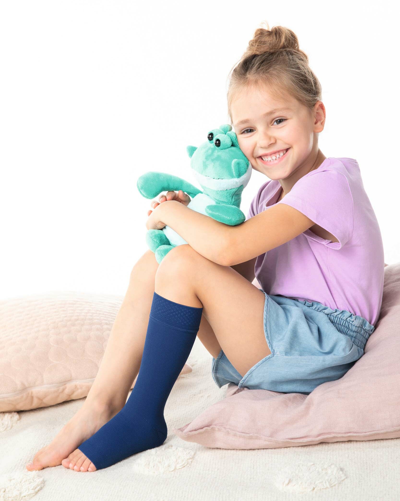 Mädchen mit Julius Kuscheltier Ein blondes Mädchen sitzt auf einem Kissen, am linken Bein eine blaue Kompressionsversorgung und hält ein Julius Kuscheltier im Arm