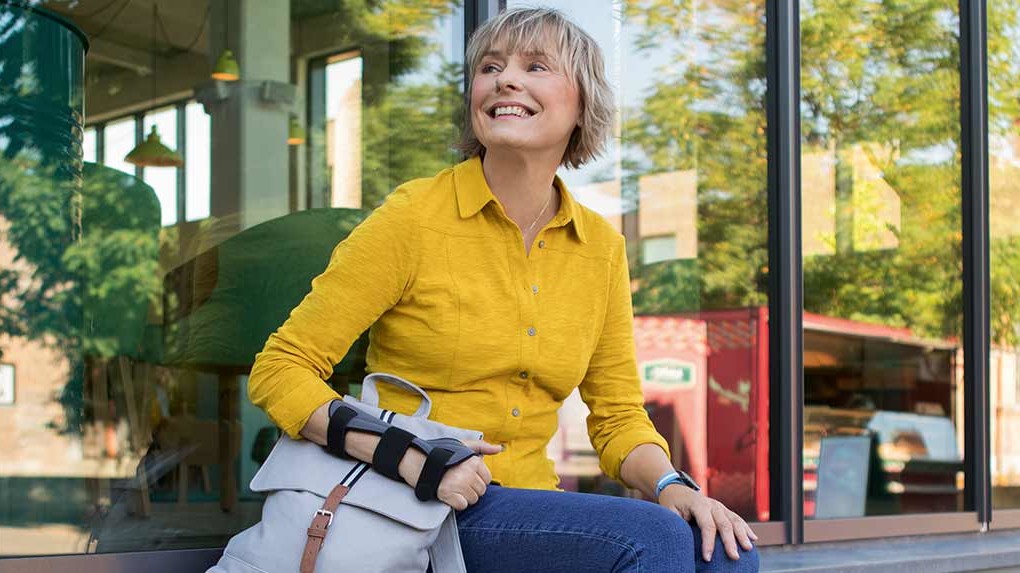 Mainteaser Ortezy Kobieta ze stabilizatorem miękkim nadgarstka JuzoPro Manu Xtec Palmar siedzi przed szklaną fasadą i patrzy z uśmiechem w dal