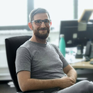 Mitarbeiter in der IT lächelt in die Kamera, während er auf einem Bürostuhl sitzt